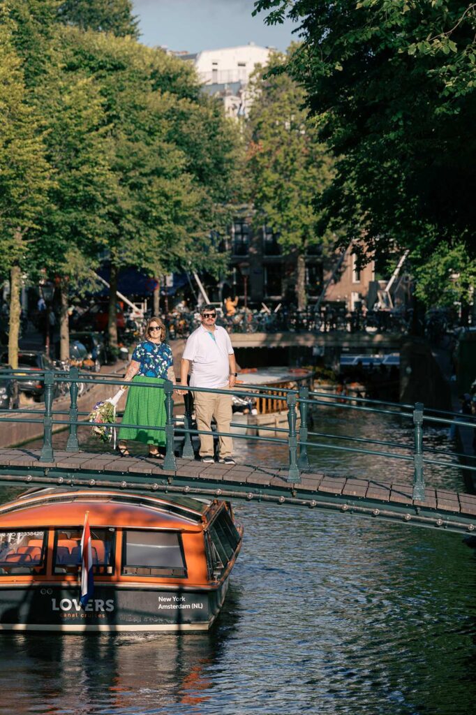 A couple celebrates their 20th anniversary in Amsterdam, standing on a bridge over a canal as a tour boat passes below.