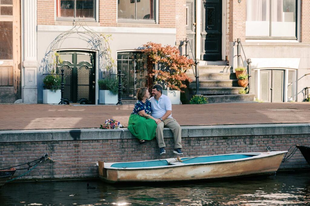 A couple sits by a canal in Amsterdam, smiling at each other, with a custom bouquet and a small boat nearby, celebrating their 20th anniversary.