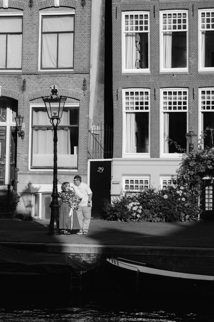 Black and white image of a couple leaning against a lampost and exchanging a look on a sunlit sidewalk in front of a brick building with large windows