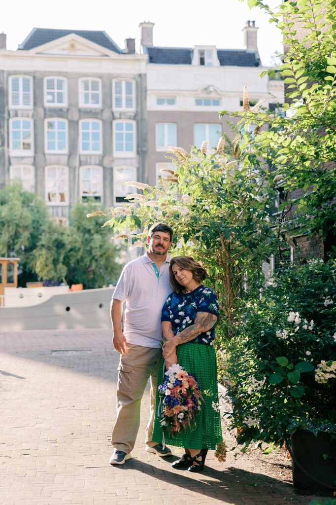 A couple poses on a sunlit Amsterdam street surrounded by greenery. Their vibe is lightly badass as the stare down the camera