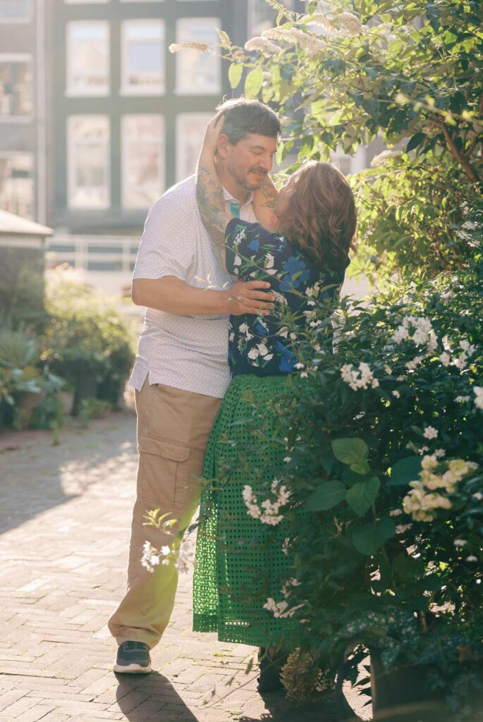 A couple gazes at each other, partially obscured by a flowering bush in the hazy golden hour light.