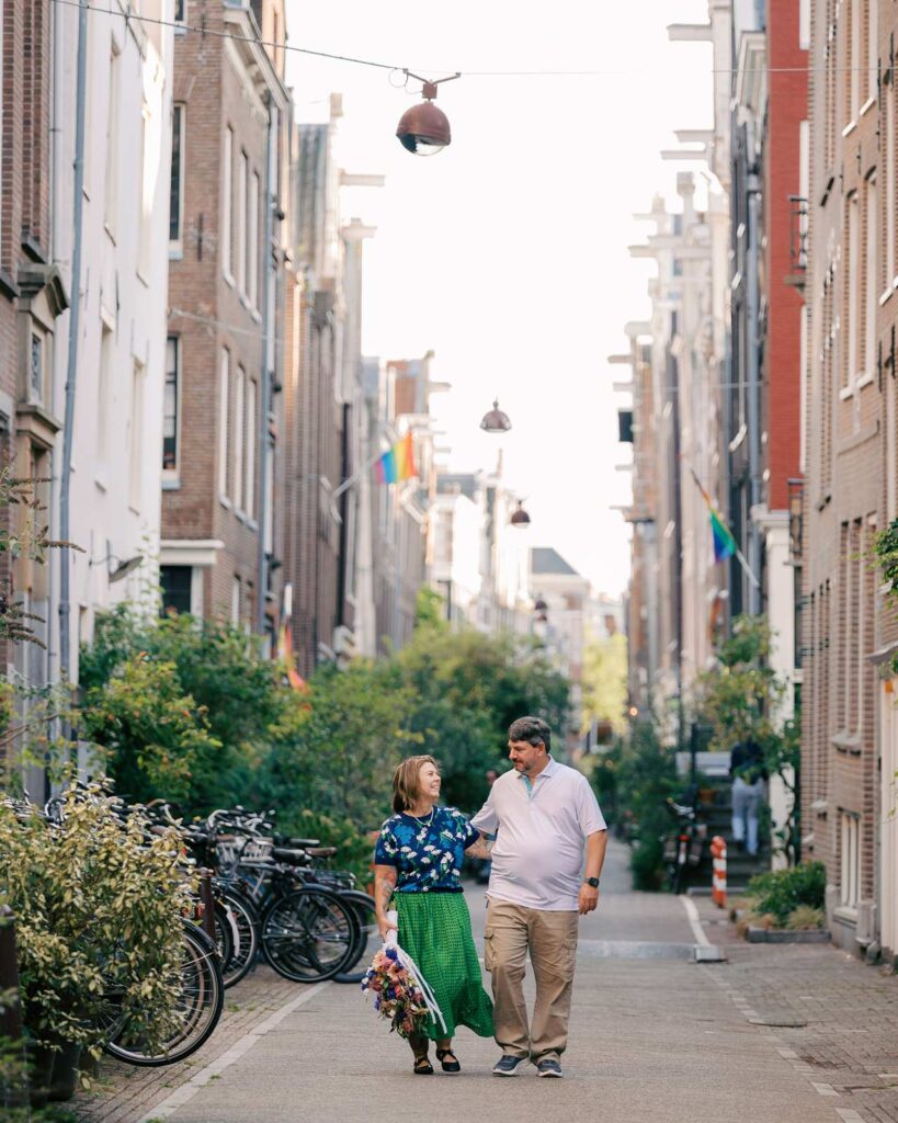 A couple walks with arms around each other an Amsterdam city street lined with bikes and plants. She is holding a colorful bouquet down by her side.