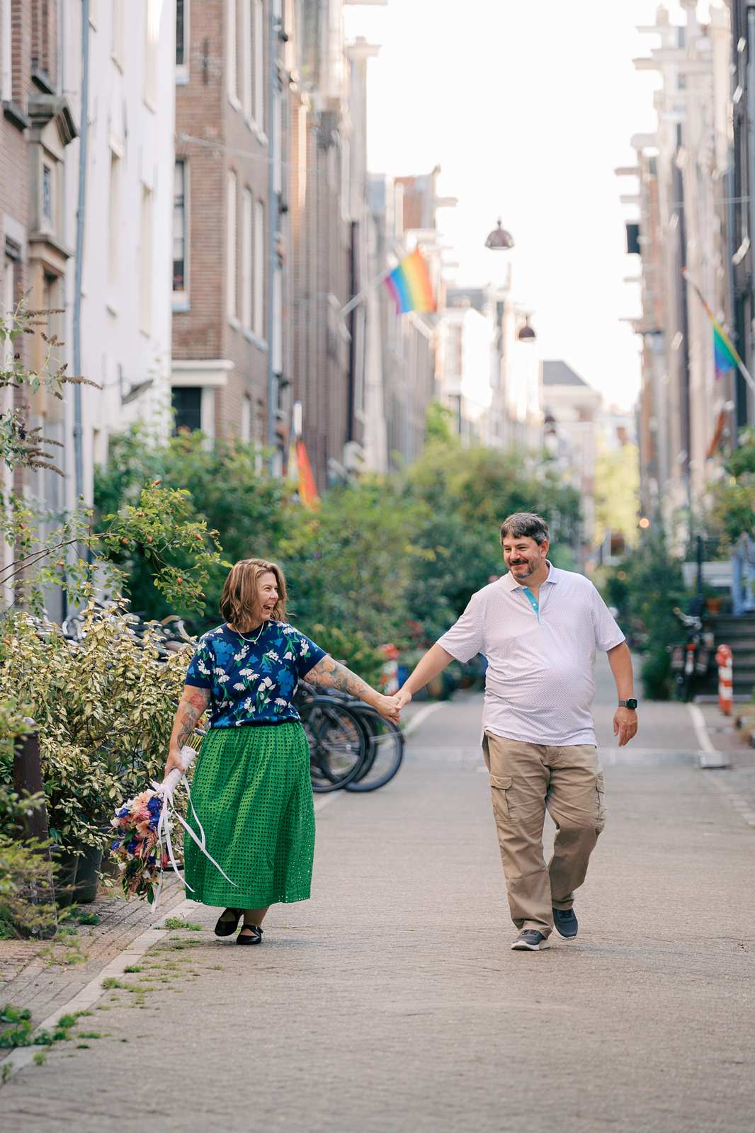 A couple holds hands and laughs while walking down an Amsterdam city street with rainbow flags, greenery, and bikes blurred in the background, during their anniversary photo shoot, complete with a custom bouquet for the occasion.