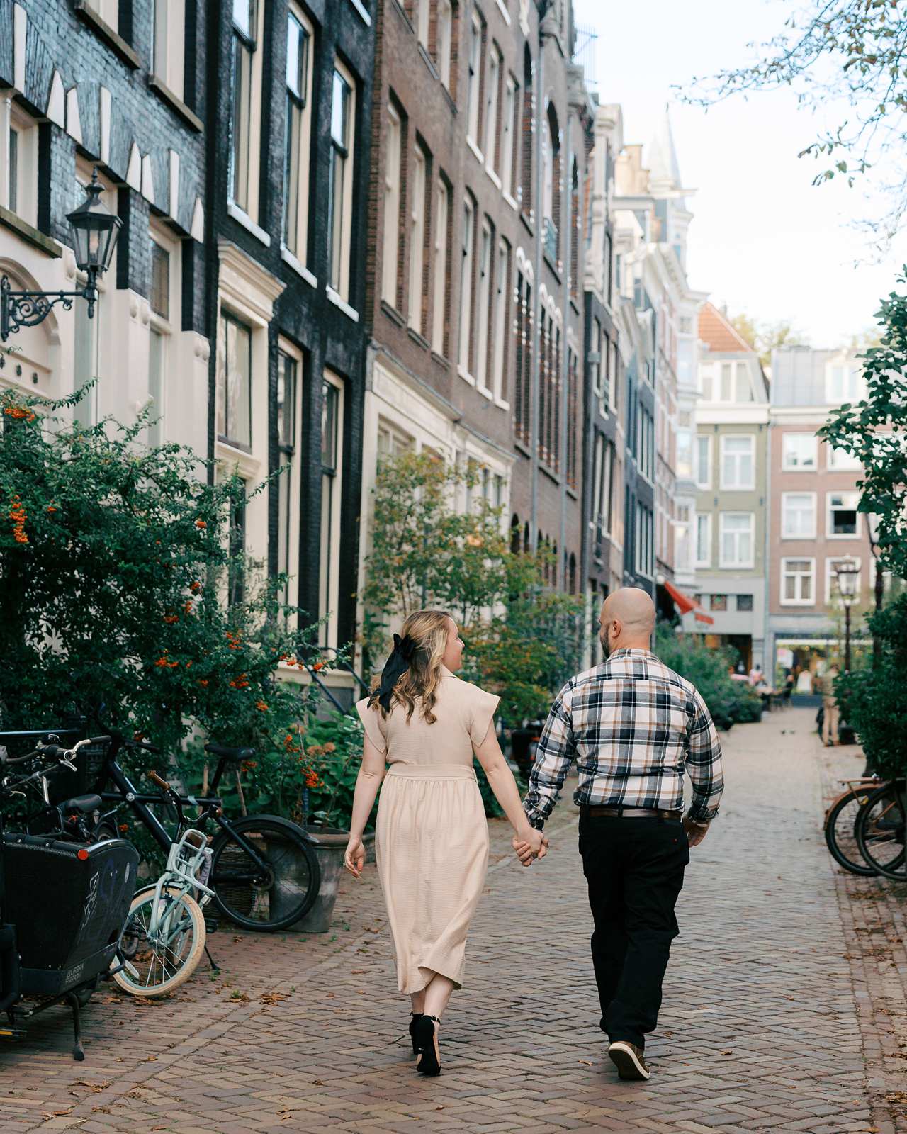 A recently engaged couple holding hands walks down a cobblestone street lined with bikes and tall buildings during golden hour.