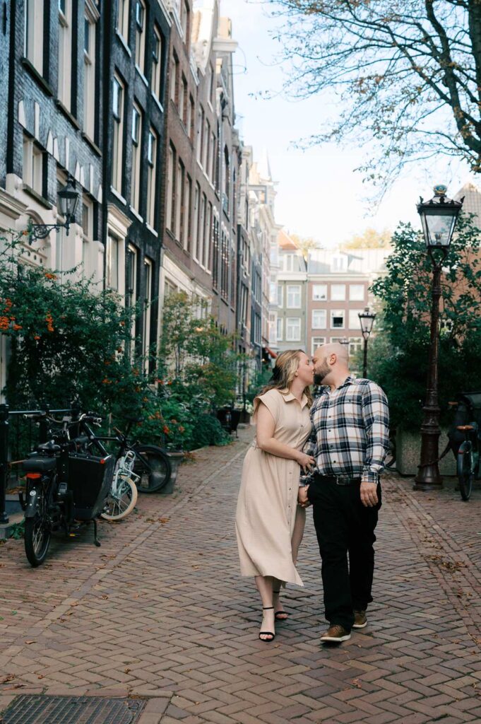 A couple kisses while walking hand in hand down a cobblestone street lined with bikes and tall buildings, basking in the golden hour glow of Amsterdam.
