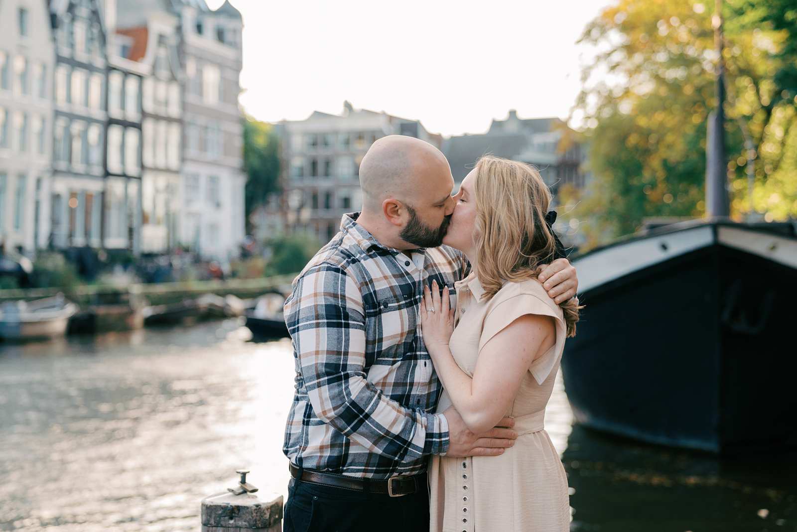 A recently engaged couple kisses by a canal in Amsterdam, with boats and historic buildings bathed in golden hour sunlight.