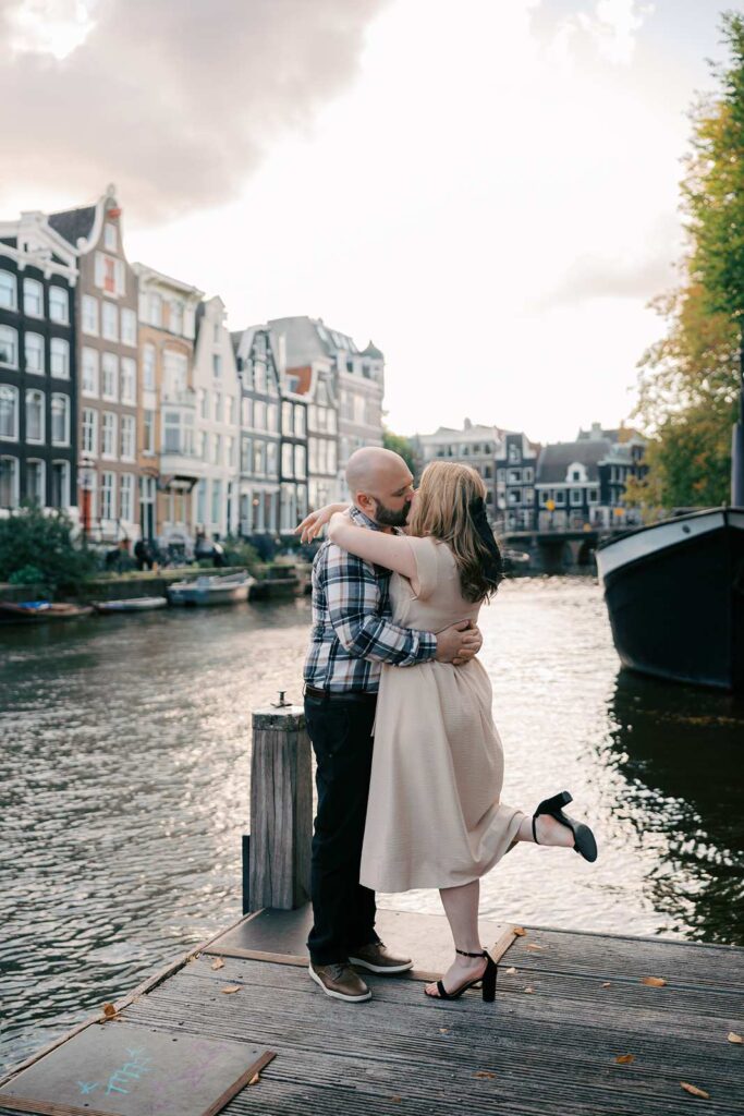 A couple embraces on a canal dock during golden hour, with classic Amsterdam houses and a boat in the background—perfect for romantic engagement photos.