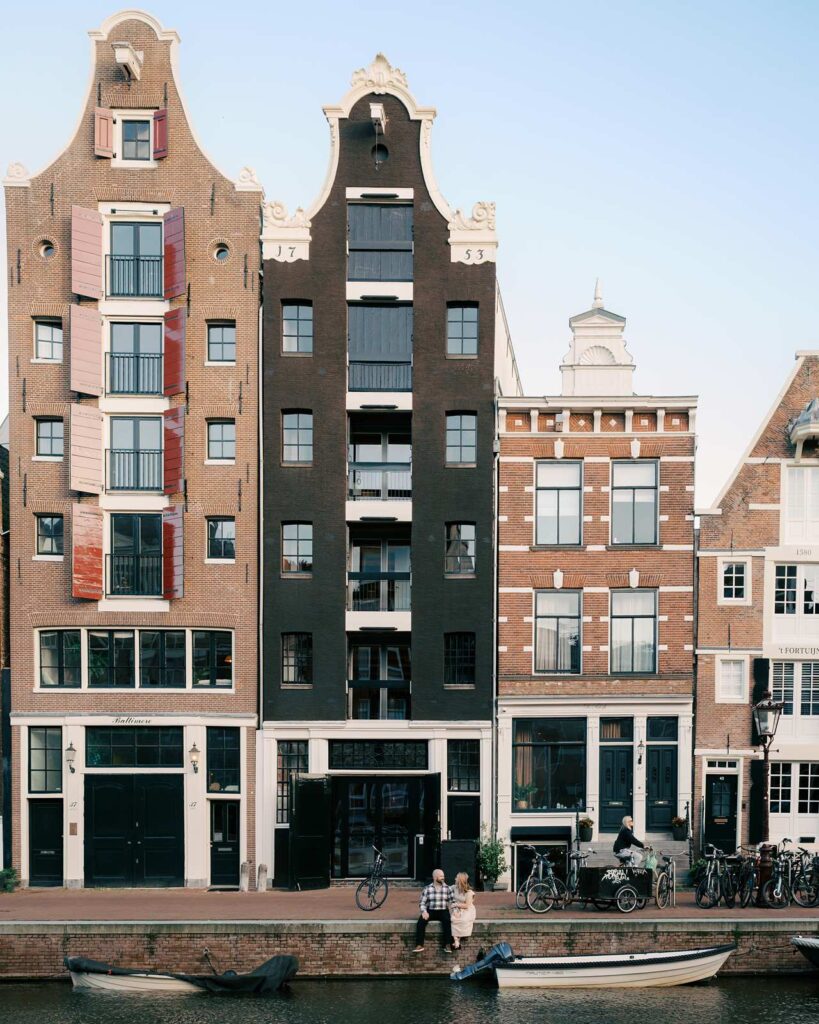 Three narrow, tall buildings face a canal in Amsterdam, where people sit and bike nearby, enjoying the golden hour—an ideal spot for engagement photos in this charming European city.
