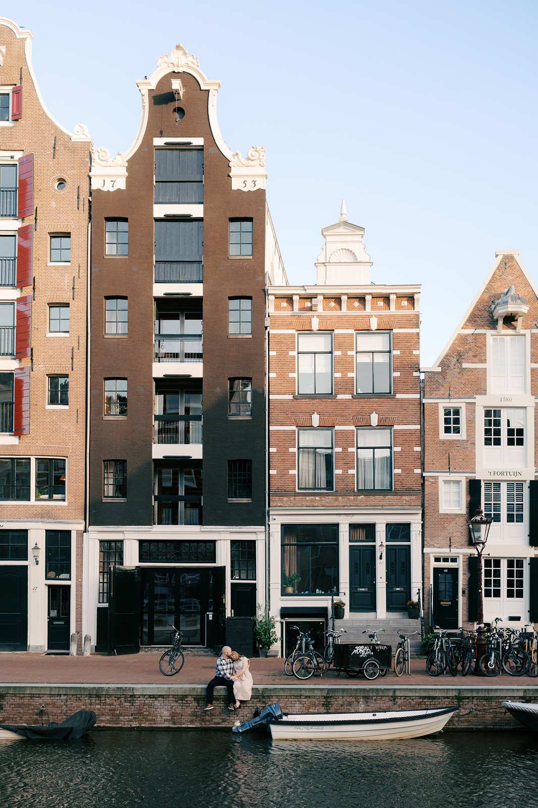 Canal-side buildings in Amsterdam with parked bicycles and people, including a recently engaged couple, sitting by the water, boat in foreground.