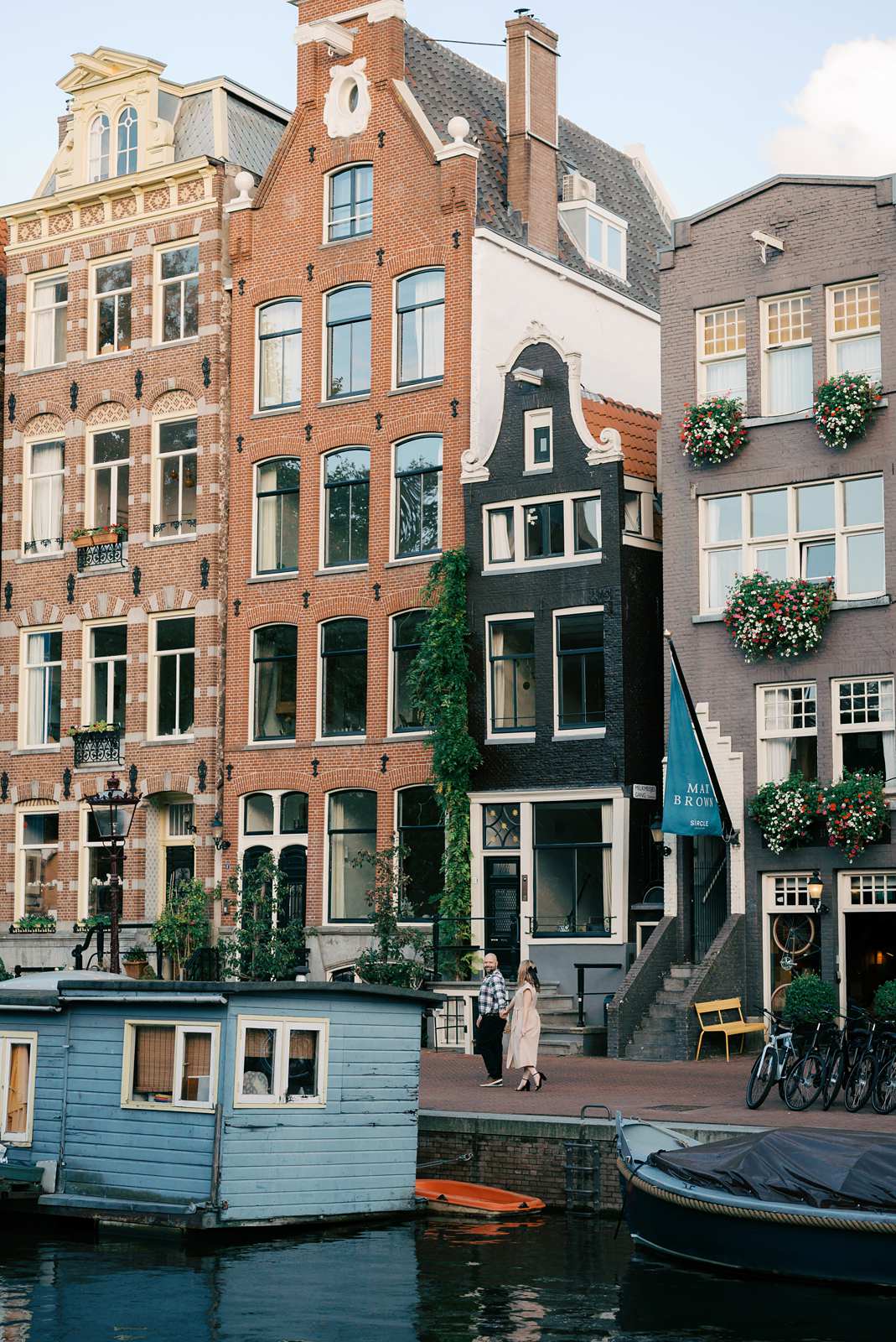 Colorful canal houses with flower boxes in Amsterdam, people walking, and a blue houseboat in the foreground, perfect for a romantic fall photo shoot during golden hour.