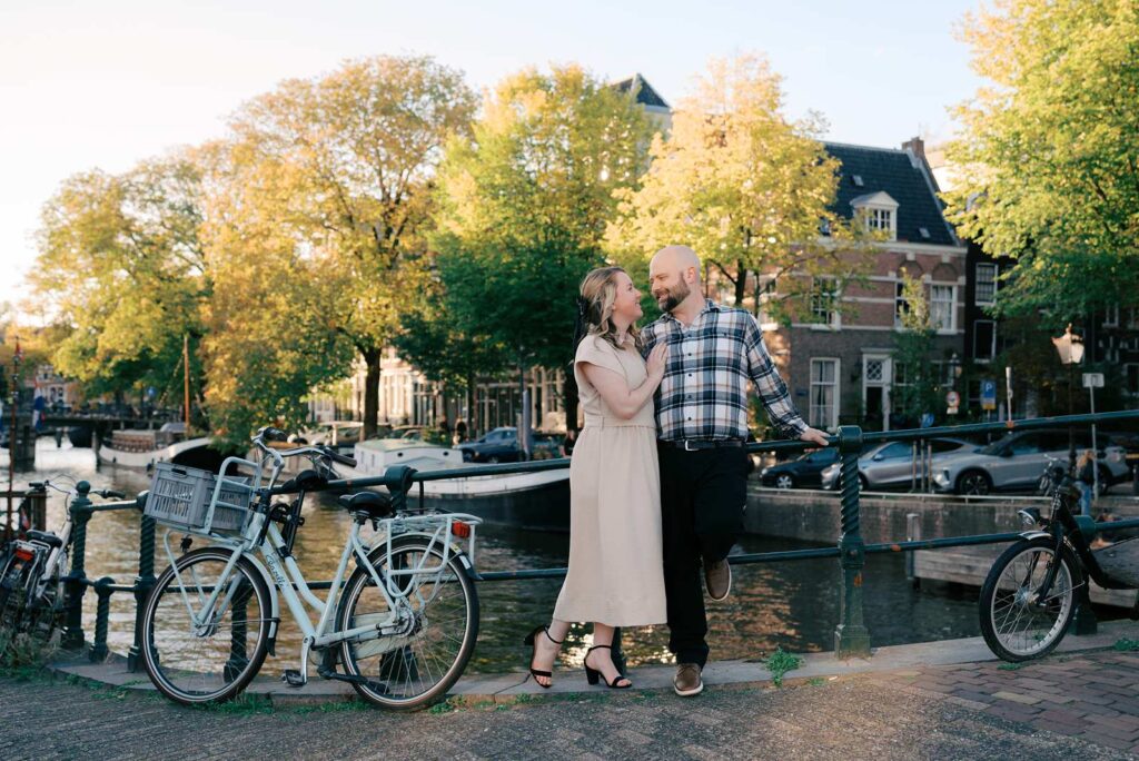 A recently engaged couple stands on a canal bridge in Amsterdam with bicycles, smiling at each other on a sunny day.