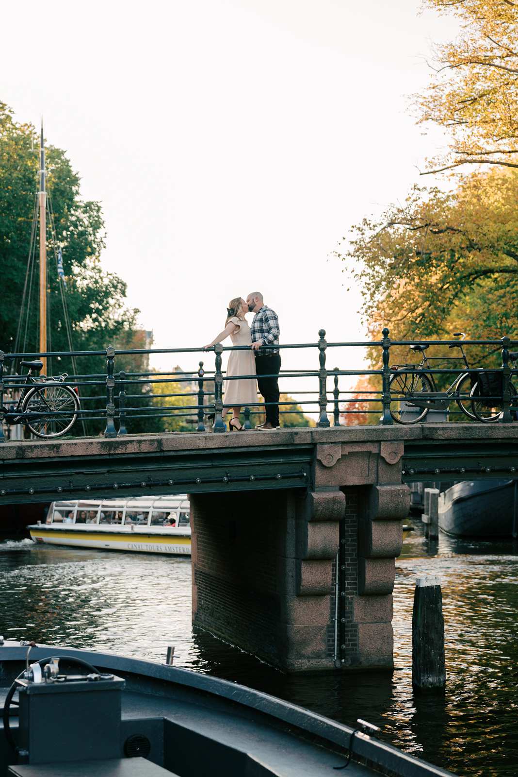 A couple stands on a bridge over a canal, sharing a kiss during golden hour, with boats and bicycles in the background—perfect for magical engagement photos.