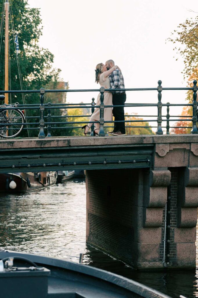 A recently engaged couple kisses on a bridge over a canal at golden hour, with trees and a bicycle in the background.