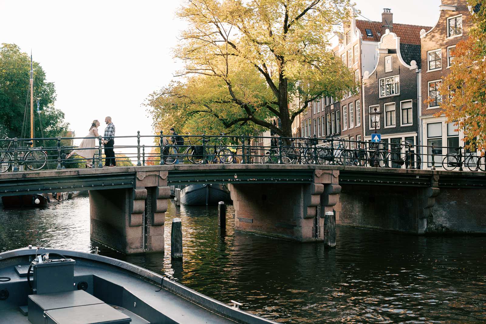 People walk and bike across a bridge over a canal lined with trees and buildings in Amsterdam, creating an ideal spot for recently engaged couples to capture memorable engagement photos.
