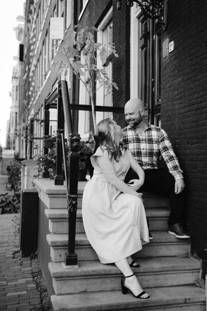 A couple sits on outdoor steps, smiling at each other in front of a brick building during a romantic fall photo shoot at golden hour.
