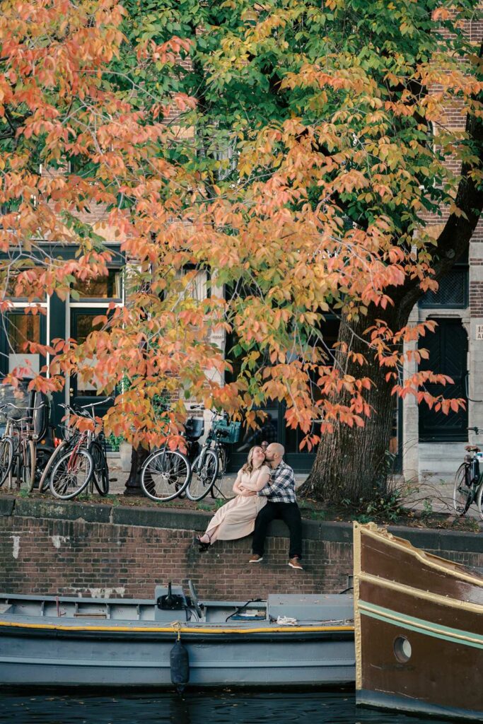 A couple sits by a canal under an autumn tree during golden hour, with bicycles lined up in the background.