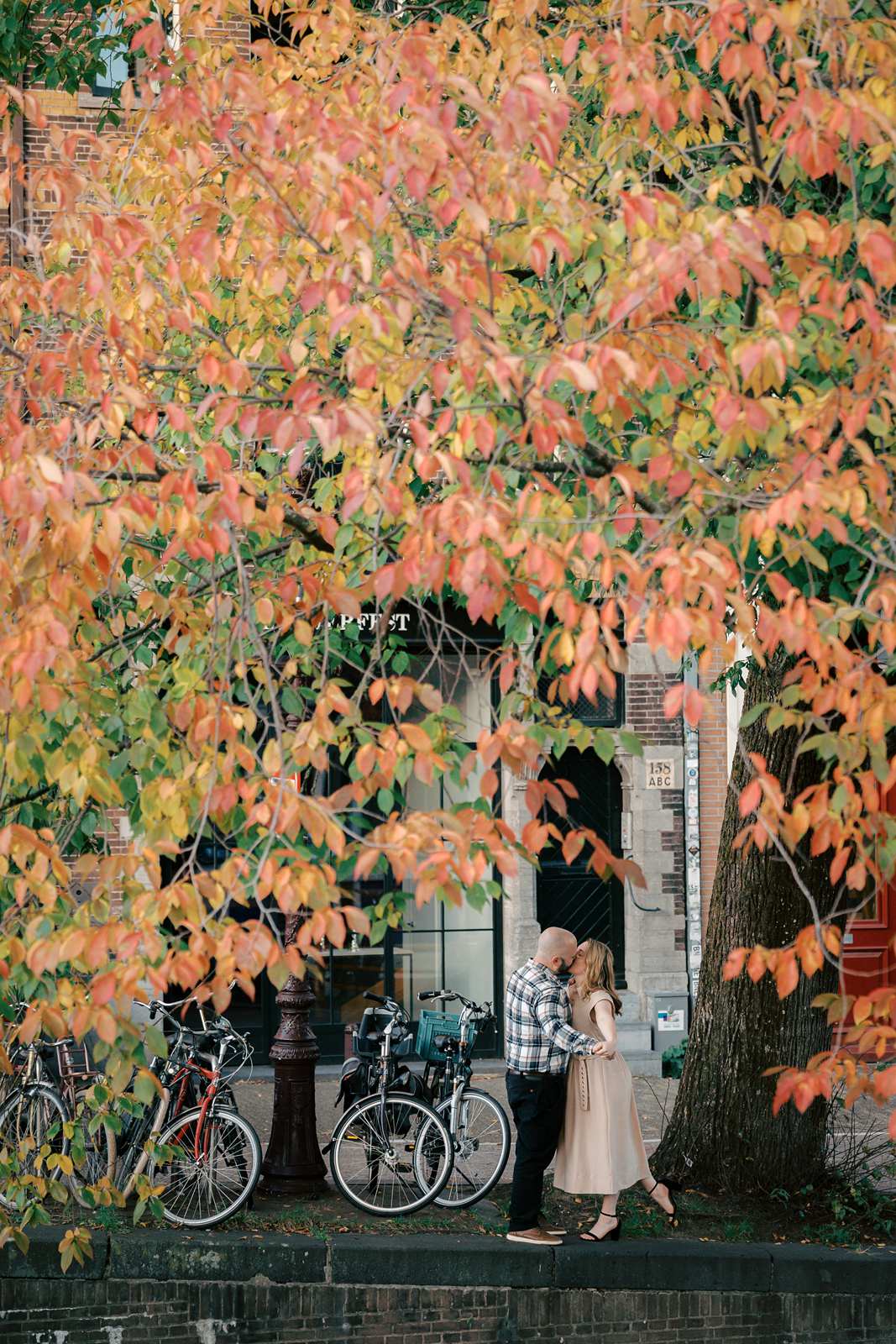 A couple embraces under a large autumn tree with orange leaves and bicycles parked nearby, creating the perfect scene for a romantic fall photo shoot at golden hour.