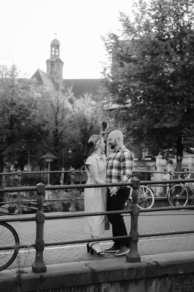 A couple stands closely on a bridge in Amsterdam during golden hour, gazing at each other with trees and a church softly glowing in the background.