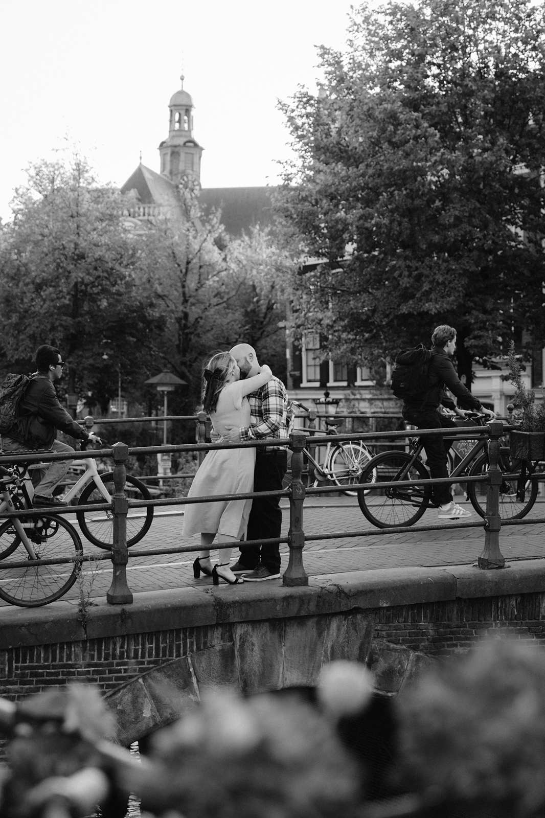 A couple kisses on a bridge as cyclists pass by, with trees and a church in the background—a perfect spot for romantic fall engagement photos.
