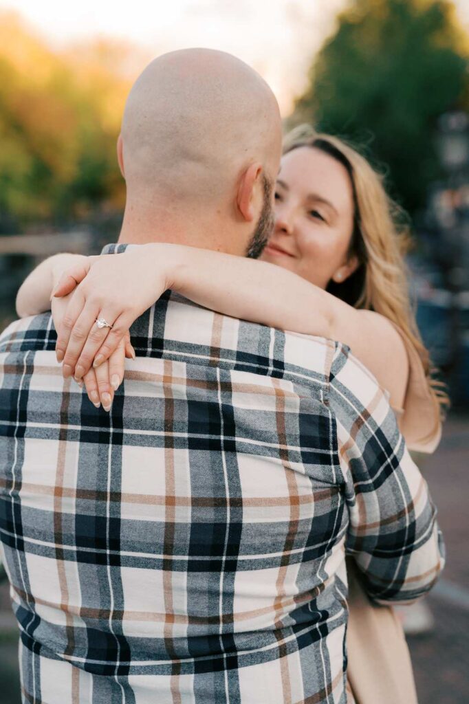 A woman hugs a man from behind, showing an engagement ring on her finger during a romantic fall photo shoot.