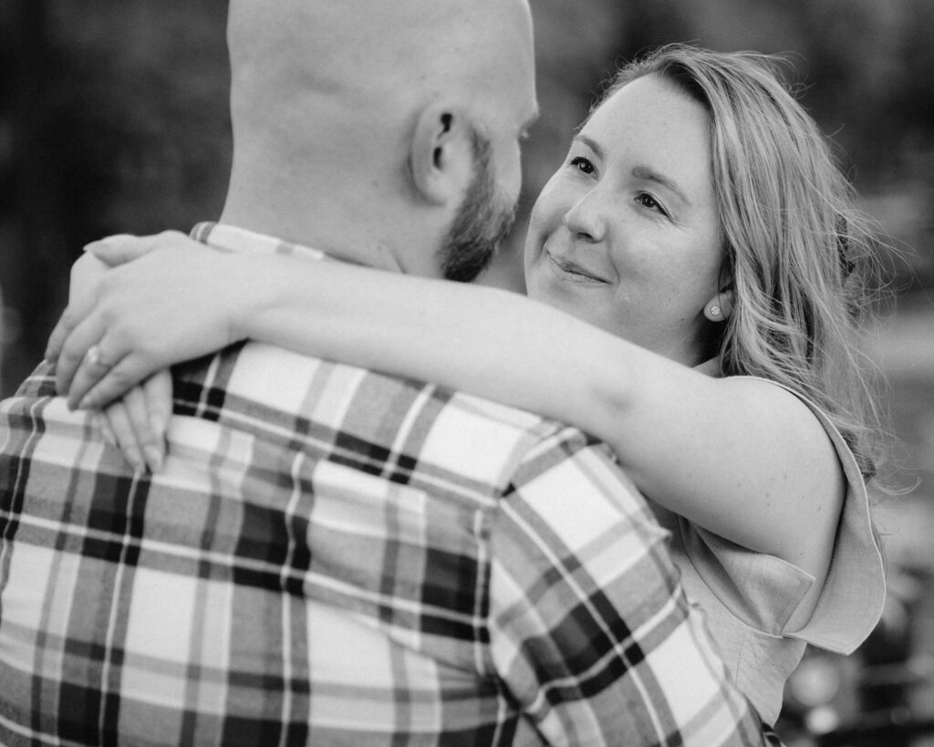 A woman smiles lovingly at a bald man as she hugs him during golden hour, both enjoying a tender moment outdoors.