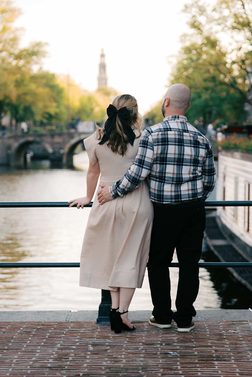 A recently engaged couple stands by a canal, embracing and looking at the water and bridge in the distance.