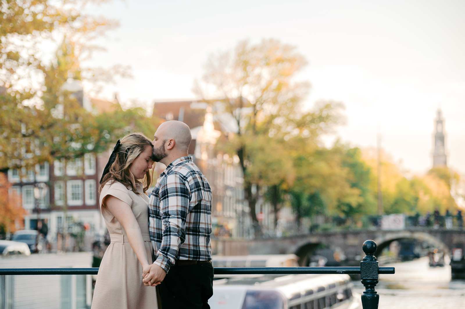 A couple stands close together, holding hands on a bridge over a canal in Amsterdam, surrounded by trees and historic buildings.