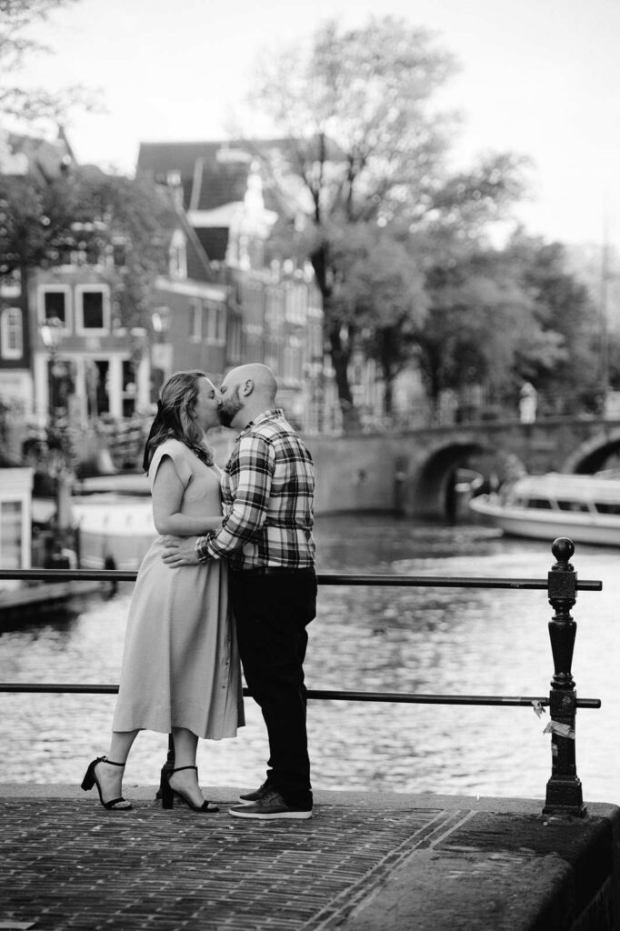 A recently engaged couple kisses by a canal in a city, with boats and houses in the background, capturing the magic of golden hour.
