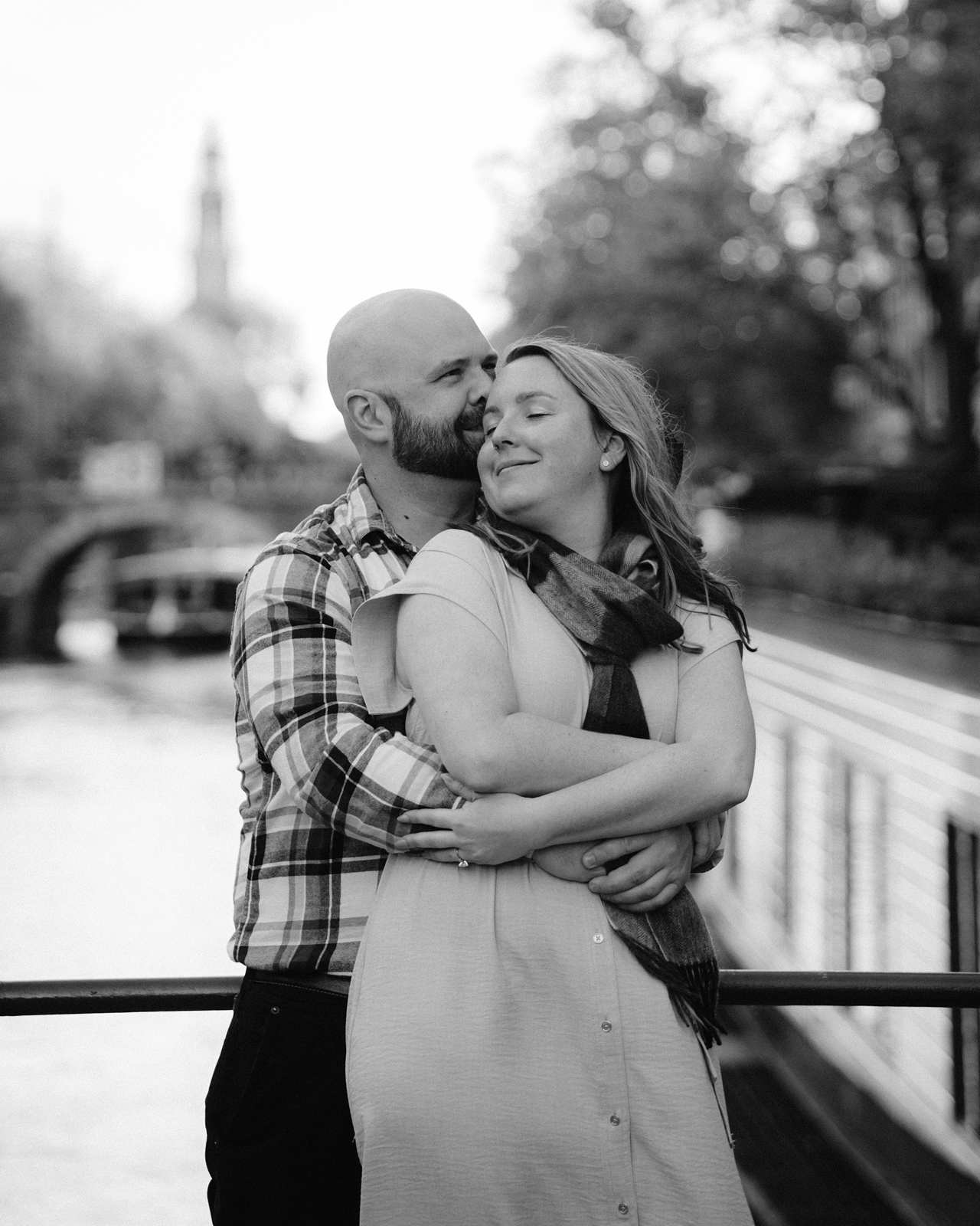 A couple embraces on a bridge in Amsterdam, smiling with eyes closed, bathed in the warm glow of golden hour in a peaceful outdoor setting.