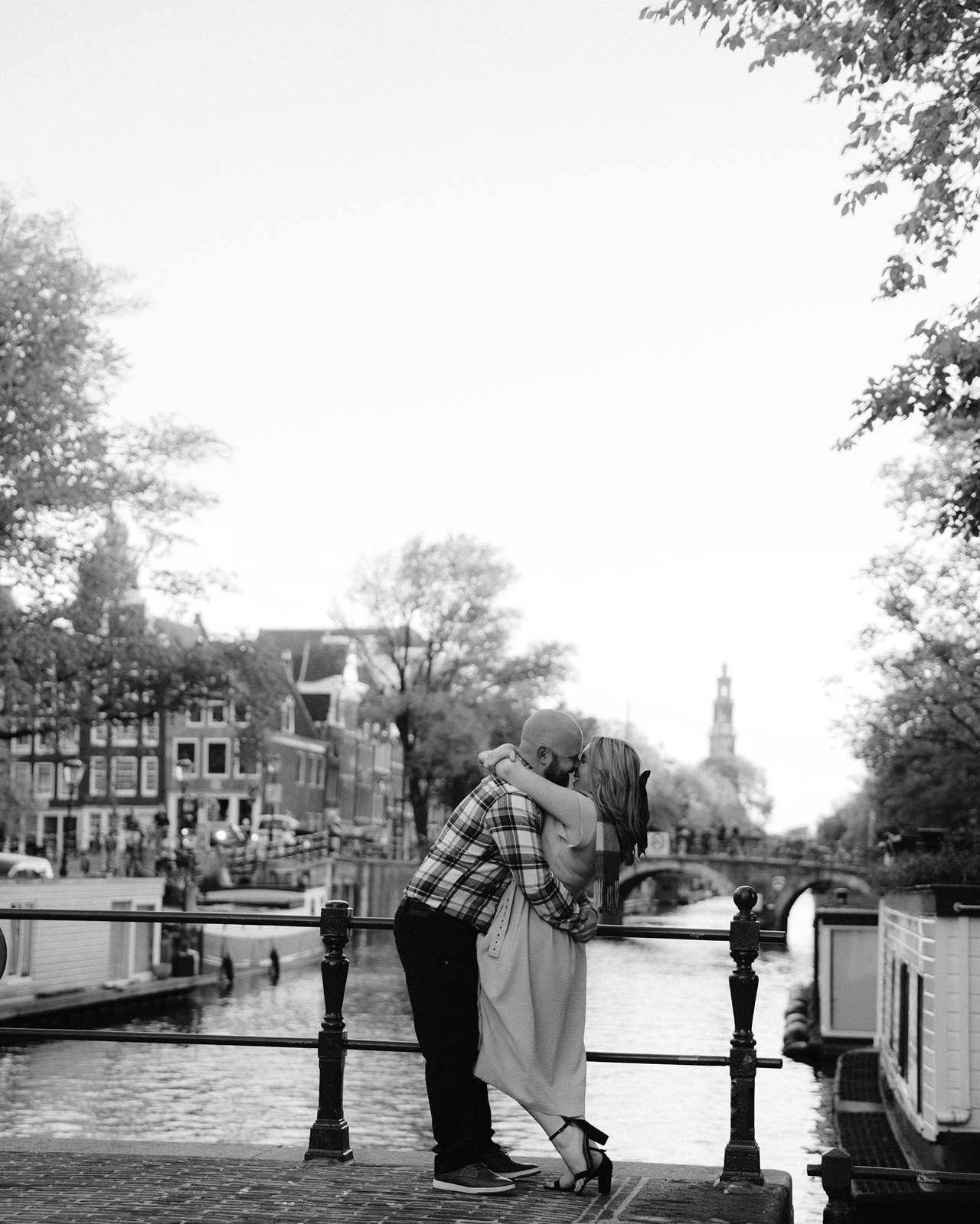 A couple, recently engaged, embraces and kisses on a canal bridge in Amsterdam, surrounded by trees and buildings.